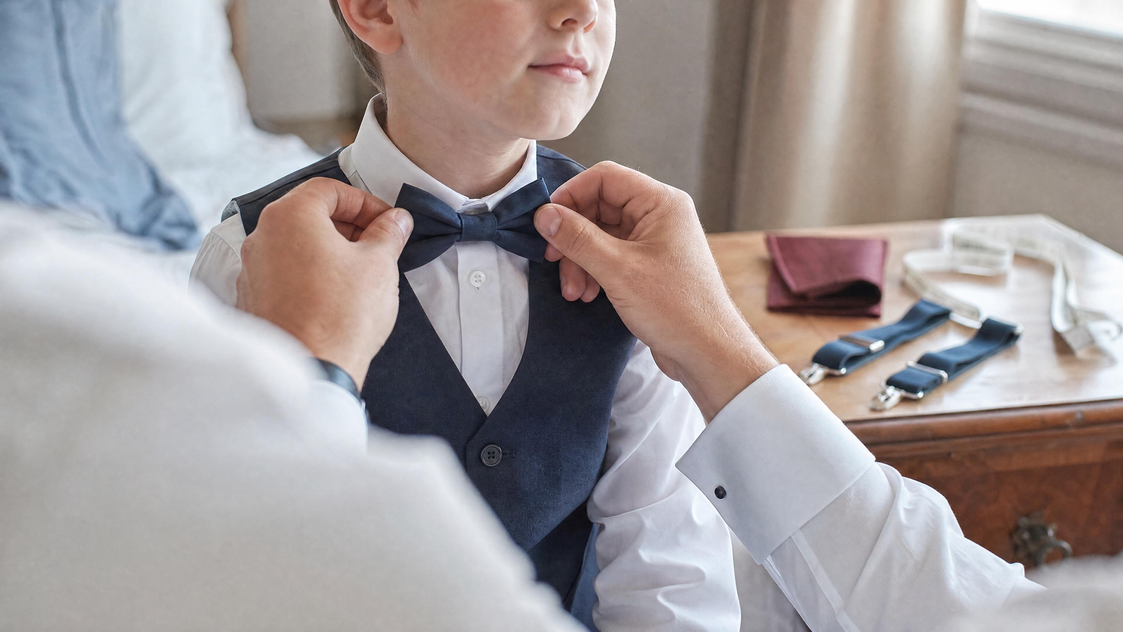 Un adulte ajuste un nœud papillon sur un enfant en chemise blanche, avec un mètre ruban visible sur une commode, dans une chambre lumineuse avant une cérémonie Obrázek